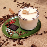 Large white coffee candle decorated with coffee beans on green ceramic plate with cinnamon sticks, star anise, and red berries on rustic burlap background