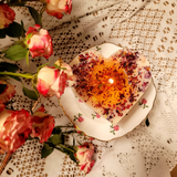 Burning Beauty Scents large heart rose petals candle with warm flame on vintage floral plate surrounded by fresh pink roses and white lace