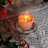 Large rose petals candle with bright burning flame illuminating dried red rose petals on surface, displayed on vintage floral china plate with gold rim, surrounded by fresh pink roses and white lace doily