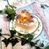 Large Heart Wild Flowers soy candle 500g burning with golden flame glow in heart-shaped wax pool, blue cornflowers visible around edges, styled on ivy-patterned plate with fresh ivy - romantic heart candle burning demonstration photography