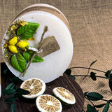 Large lemon candle with botanicals on rustic wooden board with dried citrus slices and green vine on burlap