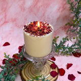 Lit medium candle topped with crushed rose petals creating warm ambiance on elegant glass stand with eucalyptus styling