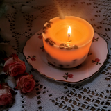 Small coffee scented candle with embedded coffee beans burning brightly on decorative rose-patterned plate with pink roses and vintage lace styling