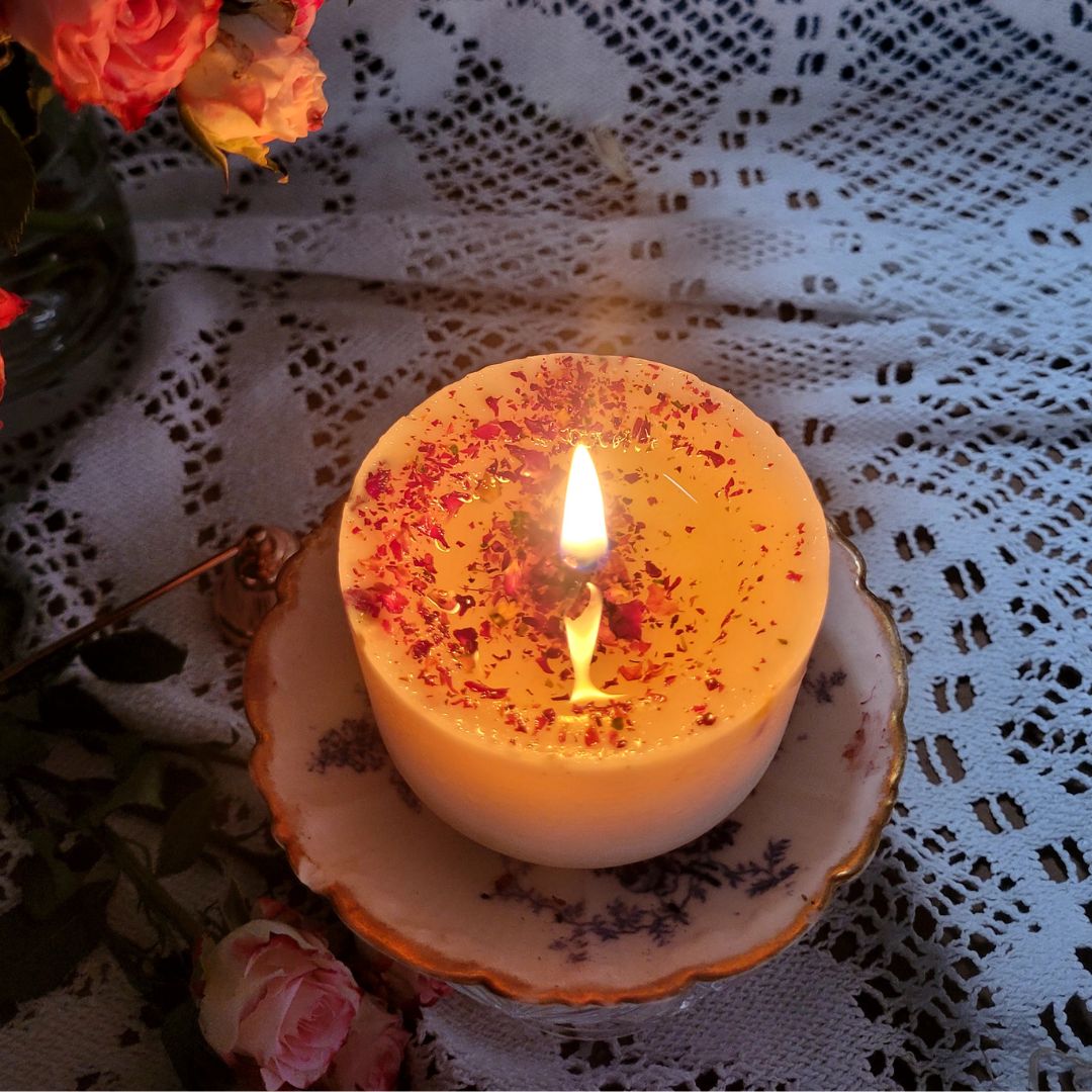 Burning small rose petals candle with warm flame on vintage plate surrounded by fresh pink roses and white lace