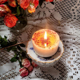 Handmade small candle with dried rose petals creating warm glow surrounded by fresh pink carnations on vintage lace