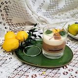 Glass candle with decorative elements on a green plate, surrounded by lemons and leaves on a lace tablecloth.