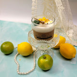 Candle with 'BS Scents' branding surrounded by lemons and limes on a light blue surface.