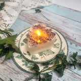 Small Heart Wild Flowers soy candle 300g burning with lit flame and melted wax pool, blue cornflowers decoration visible in heart shape, styled on ivy-patterned plate with fresh ivy - heart-shaped candle burning demonstration photography