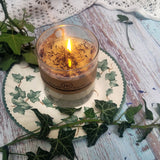 Large Glass Wild Flowers soy candle 800g burning with golden flame glow in clear glass container, blue cornflowers floating in melted wax pool, styled on ivy-patterned plate with fresh ivy - large glass container burning demonstration photography