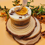 Decorative candle with an orange slice on top, branded 'Beauty Scents', on a wooden surface with foliage.