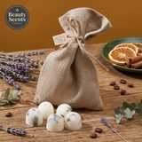 A rustic linen pouch with a twine bow and tag, branded with the Beauty Scents Candles logo, sits on a wooden table. In front of the pouch are four white botanical wax melts with visible herbs and petals. The scene is styled with sprigs of lavender, eucalyptus leaves, coffee beans, and a green plate with dried orange slices and cinnamon sticks in the background.