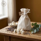 A cream linen pouch with a drawstring and branded tag sits on a rustic wooden table, surrounded by eucalyptus leaves, sprigs of lavender, and four botanical wax melts with visible herbs and petals. A bowl with more wax melts is in the background, and soft natural light enters from a nearby window. The Beauty Scents Candles logo is in the top left corner.