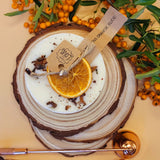 Candle with orange slice and cinnamon stick on a wooden coaster with berries and leaves.