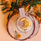 Handmade scented candle by Beauty Scents in a glass jar, topped with a dried orange slice and cinnamon sticks, displayed on rustic wood slices with orange berries and green leaves.
