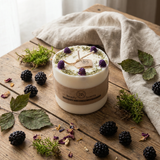 White candle with decorative elements on a wooden surface with blackberries and leaves.