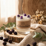Glass candle with blackberries on a wooden surface, surrounded by eucalyptus leaves and flowers.