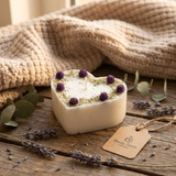 Heart-shaped candle with lavender flowers on a wooden surface with a beige blanket in the background.
