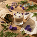 Candle with purple flowers on a wooden surface surrounded by greenery and a burlap bag.