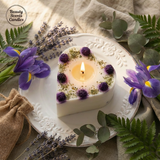 Heart-shaped candle with purple flowers on a decorative plate surrounded by greenery and lavender.