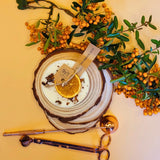 Beauty Scents candle with shredded cinnamon sticks with a wooden coaster, orange slice, and greenery on a yellow backgroundcks
