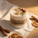 Large soy candle with wax cherries and green leaf decoration, with tag and label, styled on beige cloth on a wooden table with cinnamon sticks and dried orange slices.