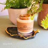 Handmade scented candle by Beauty Scents in a glass container, decorated with shredded cinnamon sticks and a dried orange slice, displayed on a dark green ceramic dish with potted ferns in the background.