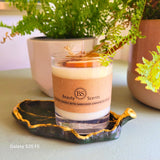 Handmade scented candle by Beauty Scents in a glass jar, decorated with shredded cinnamon sticks and a dried orange slice, placed on a green ceramic dish with potted ferns in the background.