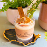 Handmade scented candle by Beauty Scents in a glass jar, decorated with a dried orange slice and cinnamon sticks, placed on a green ceramic dish with gold trim, with potted ferns in the background.