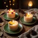 Three lit Beauty Scents small pillar candles—lemon & lemongrass, raspberry, and blueberry—displayed on green ceramic plates on a rustic wooden table with pinecones and autumn leaves, warm evening ambiance, square image.