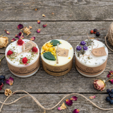 Three Beauty Scents small pillar candles—lemon & lemongrass, raspberry, and blueberry—lined up on a rustic wooden surface with scattered dried petals, berries, and twine, square image.