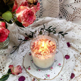 Burning Beauty Scents large glass rose petals candle with warm flame on vintage floral plate surrounded by fresh pink roses and white lace