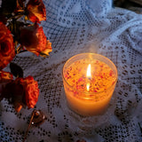 Lit large glass candle topped with crushed rose petals creating warm ambiance on vintage floral plate with fresh roses and lace doilies