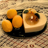 Heart-shaped candle with a lit wick on a green plate with lemons, on a lace tablecloth.