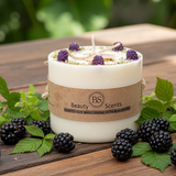 White candle with a brown label surrounded by blackberries and green leaves on a wooden surface