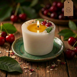 Lit large soy candle with a melted wax pool, decorated with wax cherries and green leaf detail, on a green ceramic plate on a wooden table with fresh cherries in the background.