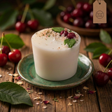 Large soy candle with wax cherries and green leaf decoration on a green ceramic plate, styled on a rustic wooden table with fresh cherries in the background.