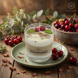 Large soy candle in a glass jar with wax cherries and green leaf decoration on a green plate on a rustic wooden table, styled next to a bowl of fresh cherries