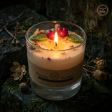 Lit large soy candle in a glass jar with a melted wax pool, decorated with wax cherries and green leaf details, styled on a dark green stone surface with dried botanicals.