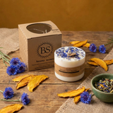 Large wildflower candle with blue petals, next to a Beauty Scents box on a rustic wooden table, styled with yellow dried leaves and blue flowers.