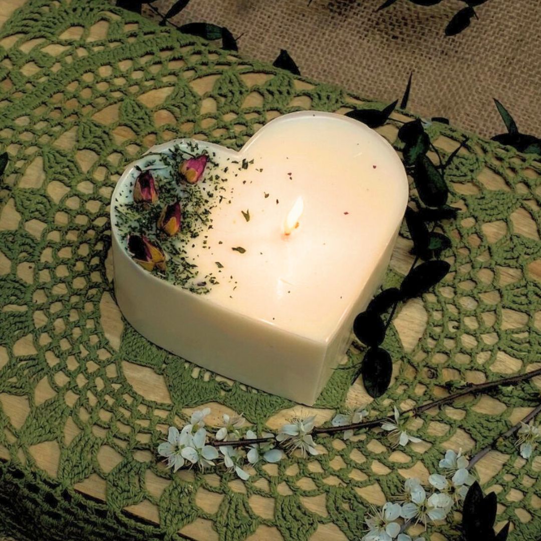 Heart-shaped candle with a lit flame on a green lace doily with flowers and leaves.