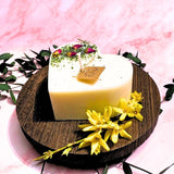White candle with a brown label on a wooden board with yellow flowers and green leaves against a pink background