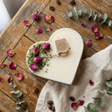 Heart-shaped candle with dried flowers and eucalyptus on a wooden surface with coffee beans and rose petals.