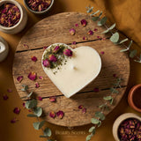 Heart-shaped candle on a wooden board with dried flowers and eucalyptus leaves, surrounded by small containers of more flowers.