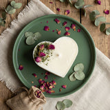 Heart-shaped candle on a green plate with dried flowers and eucalyptus leaves on a wooden surface.