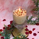 Handcrafted large glass rose petals candle with warm flame styled on crystal pedestal with eucalyptus branches and scattered petals