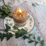 Large Glass Wild Flowers soy candle 800g burning with golden flame glow in clear glass container, blue cornflowers floating in melted wax pool, styled on ivy-patterned plate with fresh ivy - large glass container burning demonstration photography"
