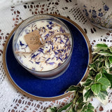 Candle in a decorative tin with dried flowers on a blue plate, surrounded by green leaves on a lace tablecloth.