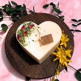 Heart-shaped candle on a wooden board with flowers and leaves on a pink background