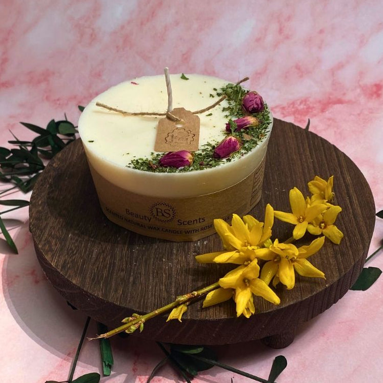A scented candle with rose buds on top, placed on a wooden surface, surrounded by decorative green leaves and a yellow flower.