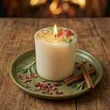 Lit medium soy candle with cherry wax melt decoration on a green ceramic plate, on a rustic wooden table with a cosy fireplace glow in the background.